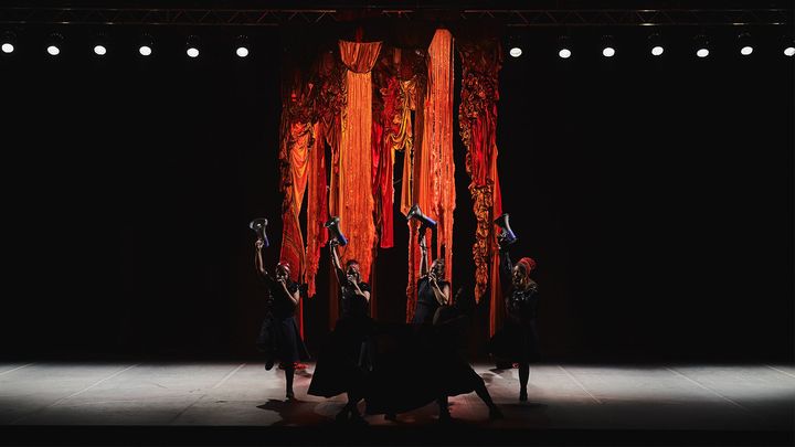 Tanssiesitys teatterilavalla, esiintyjät nostavat jalkojaan ilmassa vasten punaista taustaa.