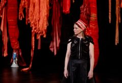 Nainen seisoo mustassa asussa teatterissa, jossa on punaisia koristeita taustalla.