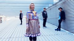 Yhtyeen jäsenet seisomassa metallisella katolla. Heistä yksi keskellä on pukeutunut koristeltuun perinteiseen asuun.