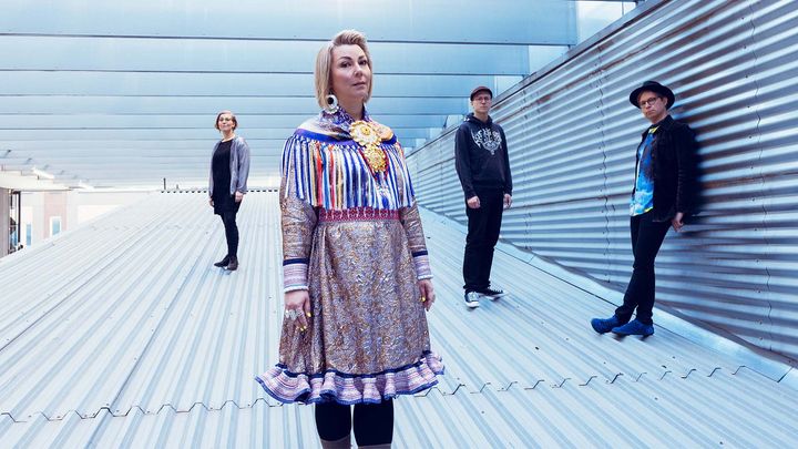 Yhtyeen jäsenet seisomassa metallisella katolla. Heistä yksi keskellä on pukeutunut koristeltuun perinteiseen asuun.