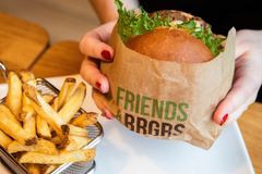 Asiakkaan kädet pitämässä hampurilaista paperipussissa; vieressä kori ranskalaisia pöydällä.