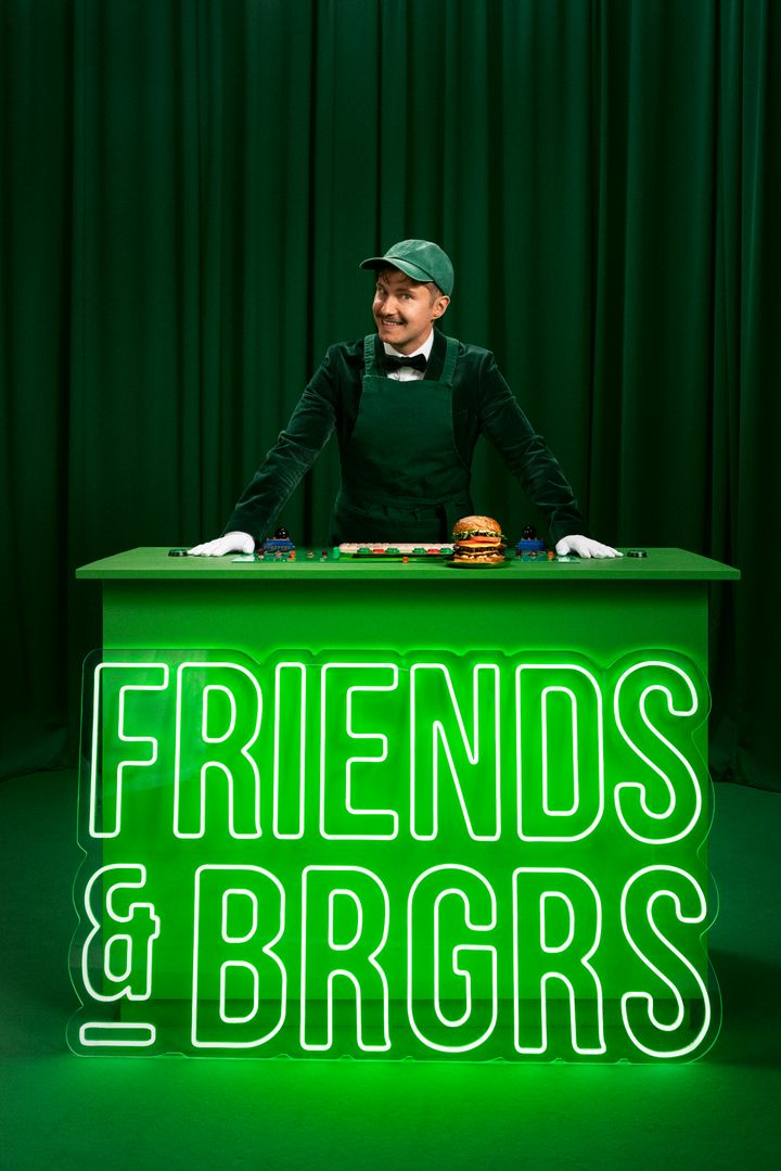 Mr. Frebu seisoo vihreän Friends & Brgrs -kontrollipöydän takana hampurilainen edessään. Taustalla vihreät verhot ja Friends & Brgrs -logo.