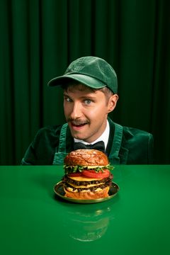 Mr. Frebu kurkistaa pöydän takaa katse kiinnittyneenä suureen hampurilaiseen vihreää taustaa vasten.