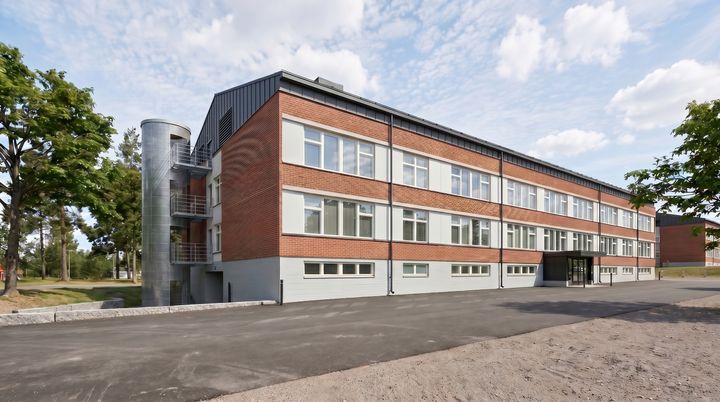Havainnekuva tiilirakennuksesta, jossa on kolme kerrosta ja rivissä isoja ikkunoita, edessä asfalttipiha ja taustalla puita.