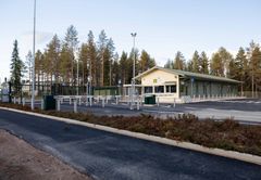 Keltainen rakennus metsäisellä alueella, edessä pysäköintialue ja puomi.