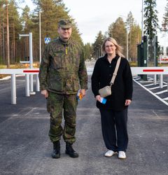 Sotilasuniformuun pukeutunut henkilö ja siviiliasuinen henkilö seisovat tien edessä, jonka taustalla on metsää ja sulkuportit.