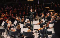 Orkesteri esiintyy lavalla konsertissa, kapellimestari johtaa muusikoita. Taustalla yleisö seuraa esitystä.