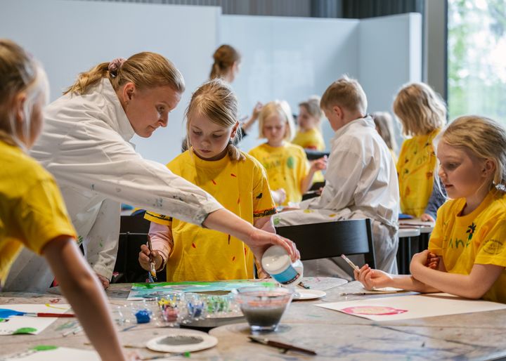 Nainen opastaa lapsia maalaustyöpajassa sisätiloissa. Lapset ovat pukeutuneet keltaisiin ja valkoisiin paitoihin.