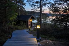 Kuvassa on kesämökin sauna järven rannalla illalla. Etualalla on valaistu polku, joka johtaa saunalle puiden keskellä.