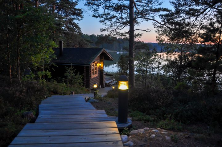 Kuvassa on kesämökin sauna järven rannalla illalla. Etualalla on valaistu polku, joka johtaa saunalle puiden keskellä.