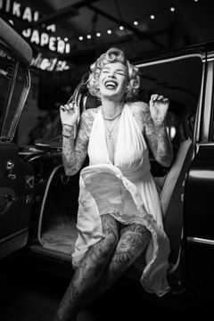 A woman with tattoos, wearing a white dress, laughing while stepping out of a vintage car.