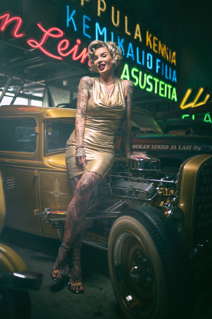 Nainen kultaisessa mekossa istuu vintage-auton konepellillä neonvalojen alla.