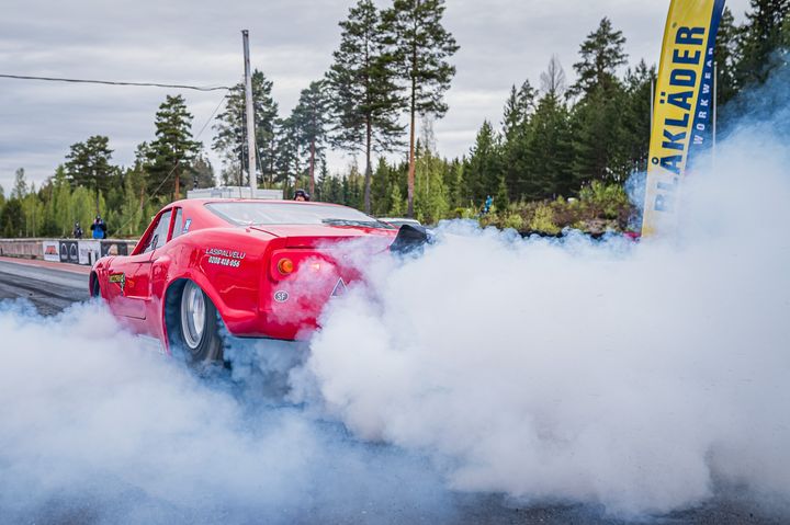 Punainen kilpa-auto aiheuttaa savua kiihdytyskilpailussa metsäisellä radalla.