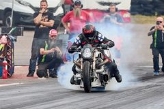 Moottoripyöräilijä lähdössä liikkeelle kiihdytysajokilpailussa, renkaista nousee savua. Taustalla ihmisiä seuraamassa.
