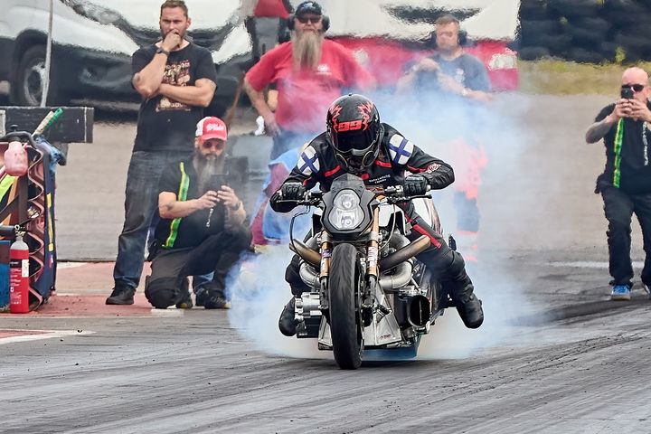 Moottoripyöräilijä lähdössä liikkeelle kiihdytysajokilpailussa, renkaista nousee savua. Taustalla ihmisiä seuraamassa.
