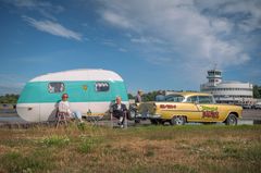 Kaksi ihmistä istuu retkituoleissa vanhan matkailuvaunun edessä, vieressä retroauto. Taustalla Helsinki-Malmin lentoasema.