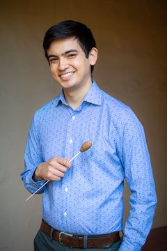 Kuvassa hymyilevä henkilö pitää kapellimestarin tahtipuikkoa kädessään. Hänellä on yllään sininen kauluspaita.