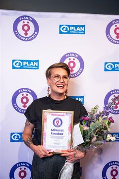 Nainen seisoo, hymyilee ja pitää käsissään palkintoa ja kukkakimppua. Taustalla on Planin tyttöpalkinnon logoja.