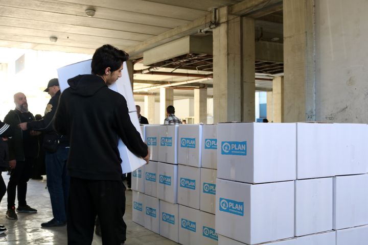 Nuori henkilö jakaa avustustarvikkeita, taustalla avustuslaatikoita Plan Internationalin logolla varustettuna.