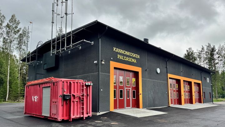 Kannuskosken paloasema. Rakennus on musta, ja siinä on punaiset ovet. Pihalla on iso punainen kontti. Taustalla näkyy metsää ja pilvinen taivas.