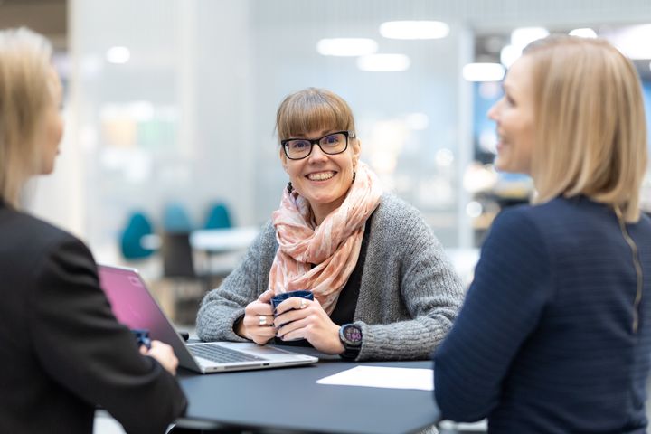 Kolme ihmistä keskustelee pöydän ääressä, yksi heistä katsoo kameraan ja hymyilee.