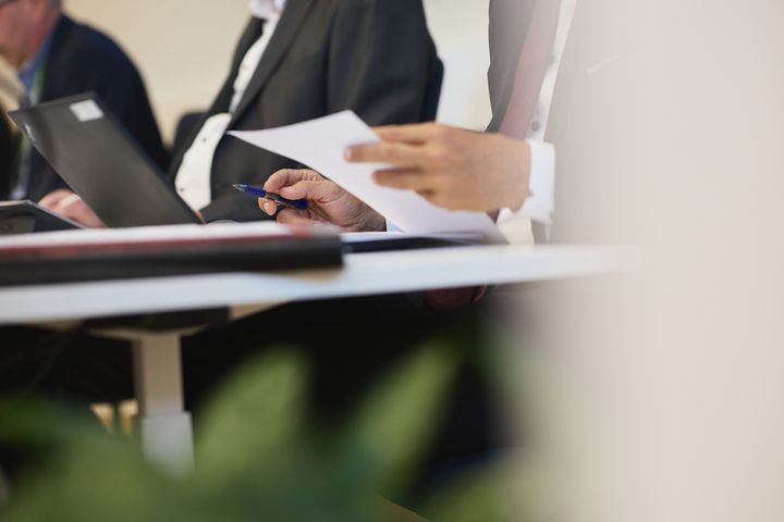 Kaksi pukuun pukeutunutta miestä pitelee papereita ja käyttää kannettavaa tietokonetta kokouksessa.