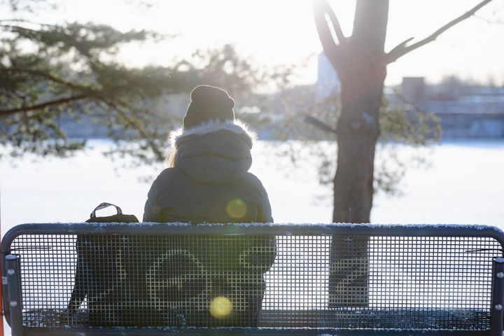 Henkilö istuu penkillä talvitakissa katsellen lumen peittämää maisemaa.