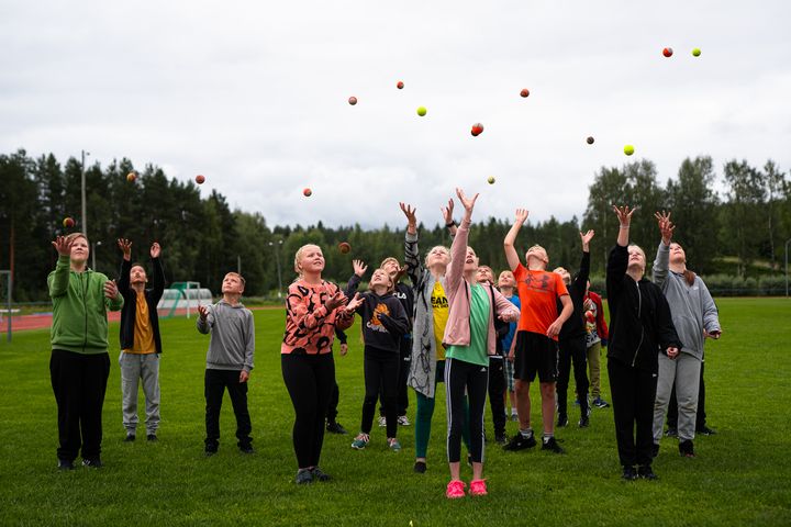 Ryhmä ihmisiä seisoo nurmikolla, heittäen palloja ilmaan. Urheilukenttä taustalla.