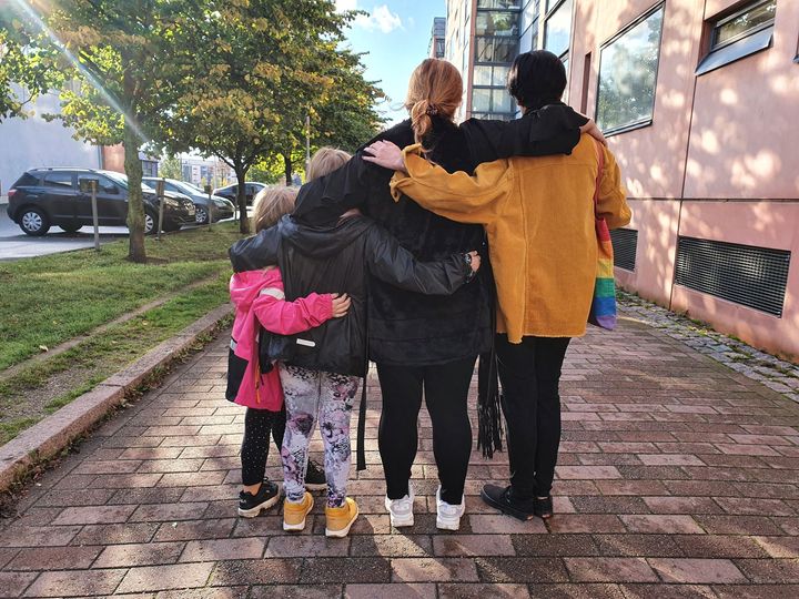 Neljä henkilöä seisoo tiellä selät kameraan päin, toisiaan halaten.