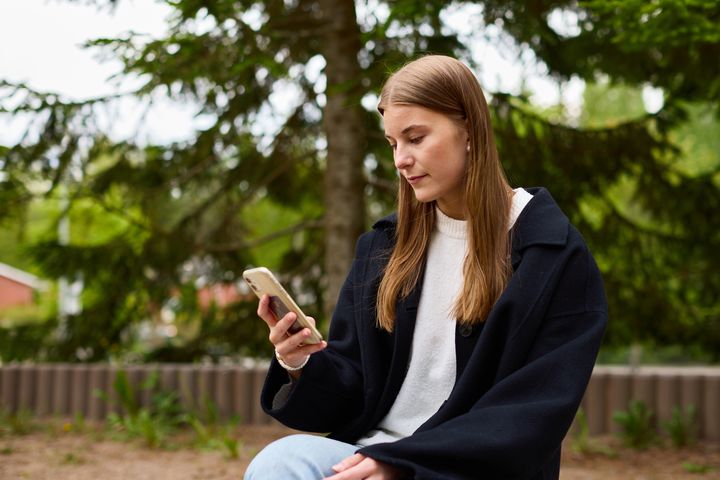Nuori nainen istuu ulkona puistossa katsellen puhelintaan.