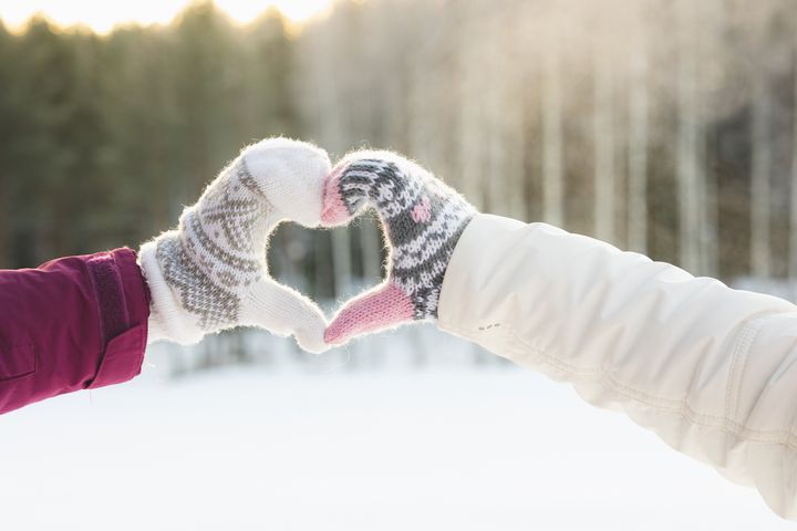 Kaksi henkilöä muodostaa käsineillään sydämen lumisella maisemalla.