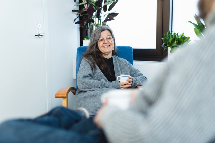 Nainen istuu tuolilla sisätiloissa ja hymyilee, kädessään kahvikuppi.