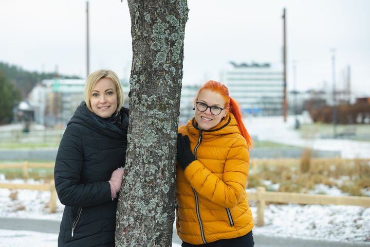 Kaksi henkilöä seisoo puun molemmin puolin talvivaatteissa.