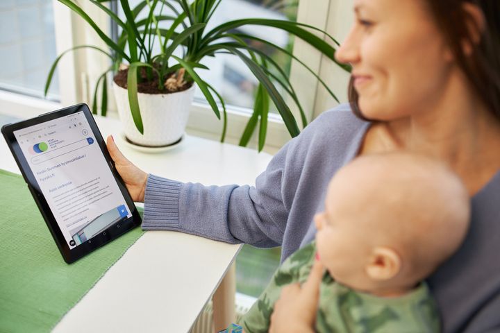 Nainen istuu ikkunan ääressä pitäen vauvaa sylissään ja katsoo tablettia.