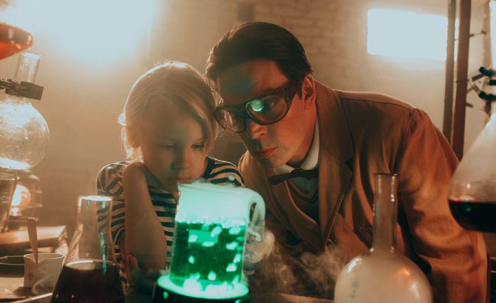 Kuvassa poika ja mies seisovat laboratoriomaisessa ympäristössä erilaisten lasisten kemiallisten välineiden keskellä. Etualalla oleva kannu hehkuu kirkkaan vihreää valoa ja sen ympärillä leijailee höyryä. Taustalla näkyy lämmintä, pehmeää valoa ja tiiliseinä, jotka luovat tunnelmallisen ja hieman mystisen ilmapiirin.