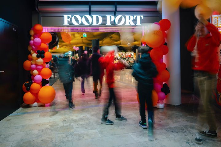 Alt-teksti: Ihmisiä kävelee sisään Redin Food Portin sisäänkäynnistä, joka on koristeltu värikkäillä ilmapalloilla.