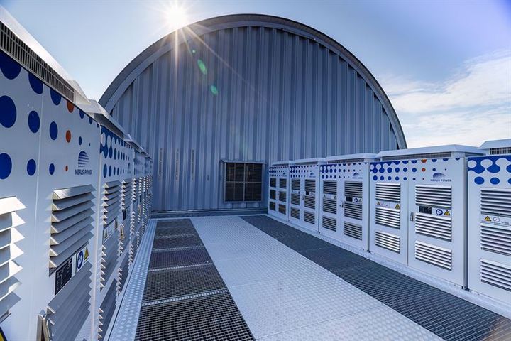 Battery storage units positioned on a rooftop with a curved metal building and the sun shining above.