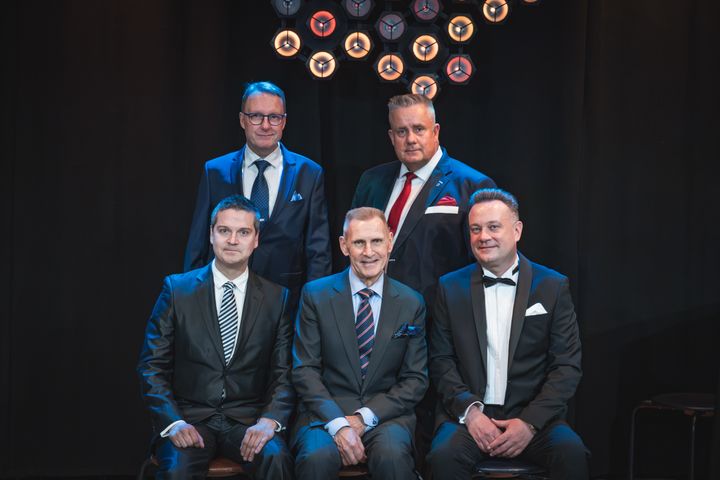 Viisi miestä poseeraa yhdessä muodollisissa puvuissa. Taustalla on tummat verhot ja ympyränmuotoisia valaisimia.