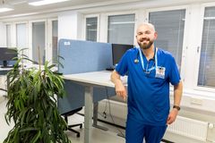 Kuvassa lääkäri sinisessä työasussa seisoo toimistossa, jossa on tietokoneita ja kasvi.