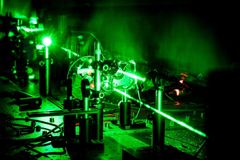 Kuva tieteellisestä laboratoriosta, jossa vihreä lasersäde kulkee optisten laitteiden läpi. Kuva on muuten pimeä, mutta lasersäde luo kuvaan vihreän hehkun.
