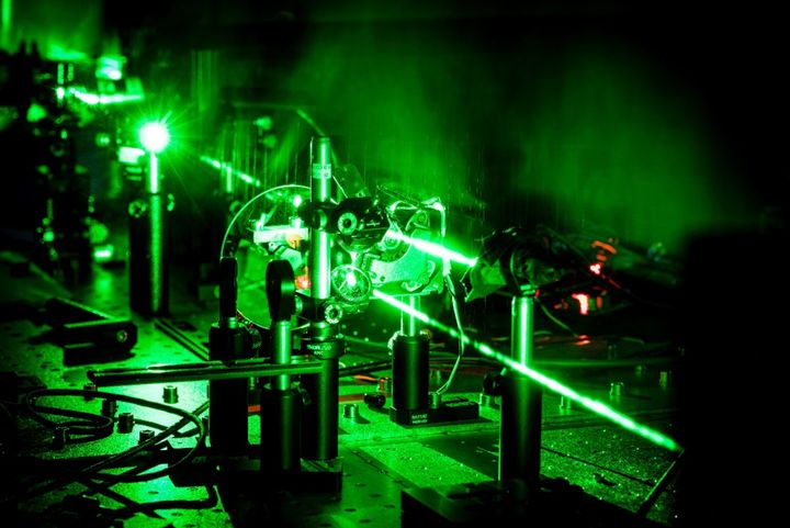 Kuva tieteellisestä laboratoriosta, jossa vihreä lasersäde kulkee optisten laitteiden läpi. Kuva on muuten pimeä, mutta lasersäde luo kuvaan vihreän hehkun.
