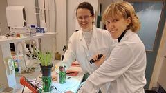Kaksi henkilöä laboratoriossa valkotakit päällä. Pöydällä heidän edessään on kaksi vihreää ainetta sisältävää lasipurkkia, joihin menee johdot.