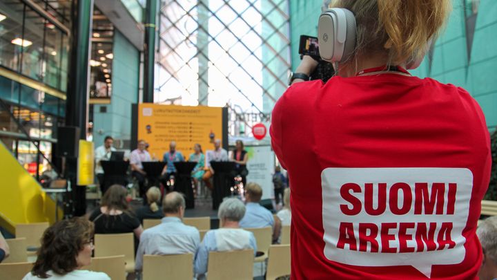 SuomiAreenan yleisöä kuvattuna takaapäin ja videokuvaaja päällään SuomiAreena-logolla varustettu paita.