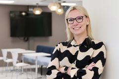 Kuva esittää hymyilevää henkilöä sisätiloissa. Hänellä on silmälasit ja vaalea tukka, ja hän nojaa vaaleaan seinään. Taustalla näkyy musta televisioruutu ja pöytiä tuoleineen.