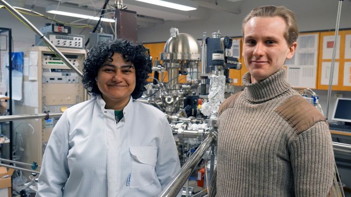 Kaksi tutkijaa vierekkäin laboratoriossa, katsovat kohti kameraa, toisella valkoinen laboratoriotakki päällä. Taustalla näkyy suurikokoinen metallinen ultrasuurtyhjiölaitteisto ja siitä moniin eri suuntiin lähteviä metalliputkia, johtoja jne.