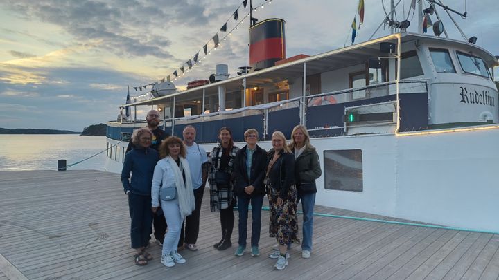 Kahdeksan henkilöä seisoo laiturilla laivan edessä.