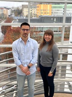 Kaksi henkilöä seisoo yhdessä sisätiloissa, heistä toinen vasemmalla ja toinen oikealla. Taustalla näkyy suuret ikkunat ja kaupunkimaisemaa.