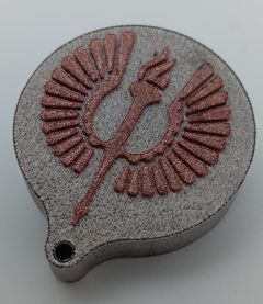 Kuvassa on metallinen harmaa kappale, jonka päällä on kohollaan kuparinvärinen Turun yliopiston siipisoihtulogo. Kappale ja logo on valmistettu metallin 3D-tulostuksella.