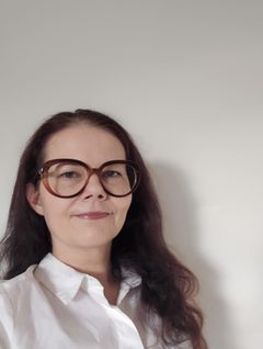 Profiilikuva väitöskirjatutkija Aleksandra Karimaasta. Henkilöllä on pitkät tummat hiukset ja suuret silmälasit ja yllään valkoinen kauluspaita. Kuvan taustalla on valkoinen seinä.