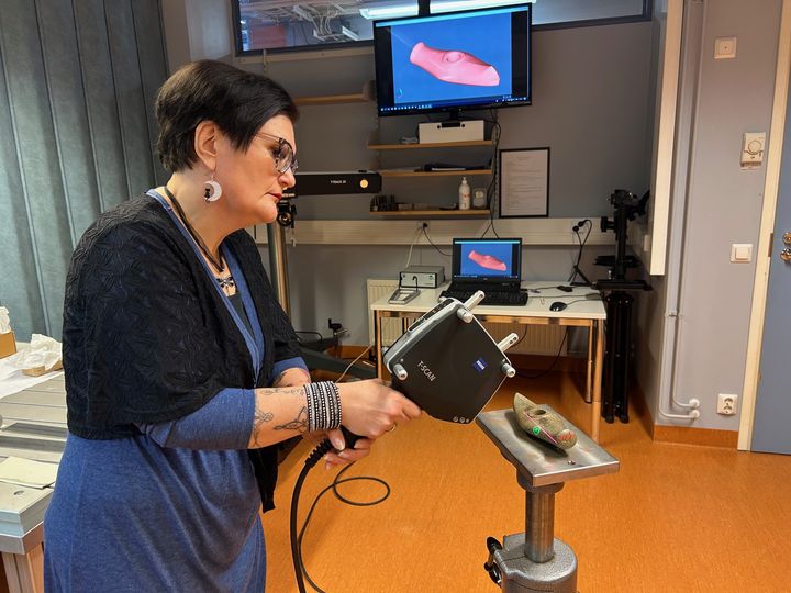 Tutkija pitää digitointilaitetta käsissään ja osoittaa sillä alustan päällä olevaa historiallista vasarakirvestä. Taustalla näkyy läppärin näyttö ja suurempi seinällä oleva näyttö, jonne digitoitu kuva on tullut näkyviin